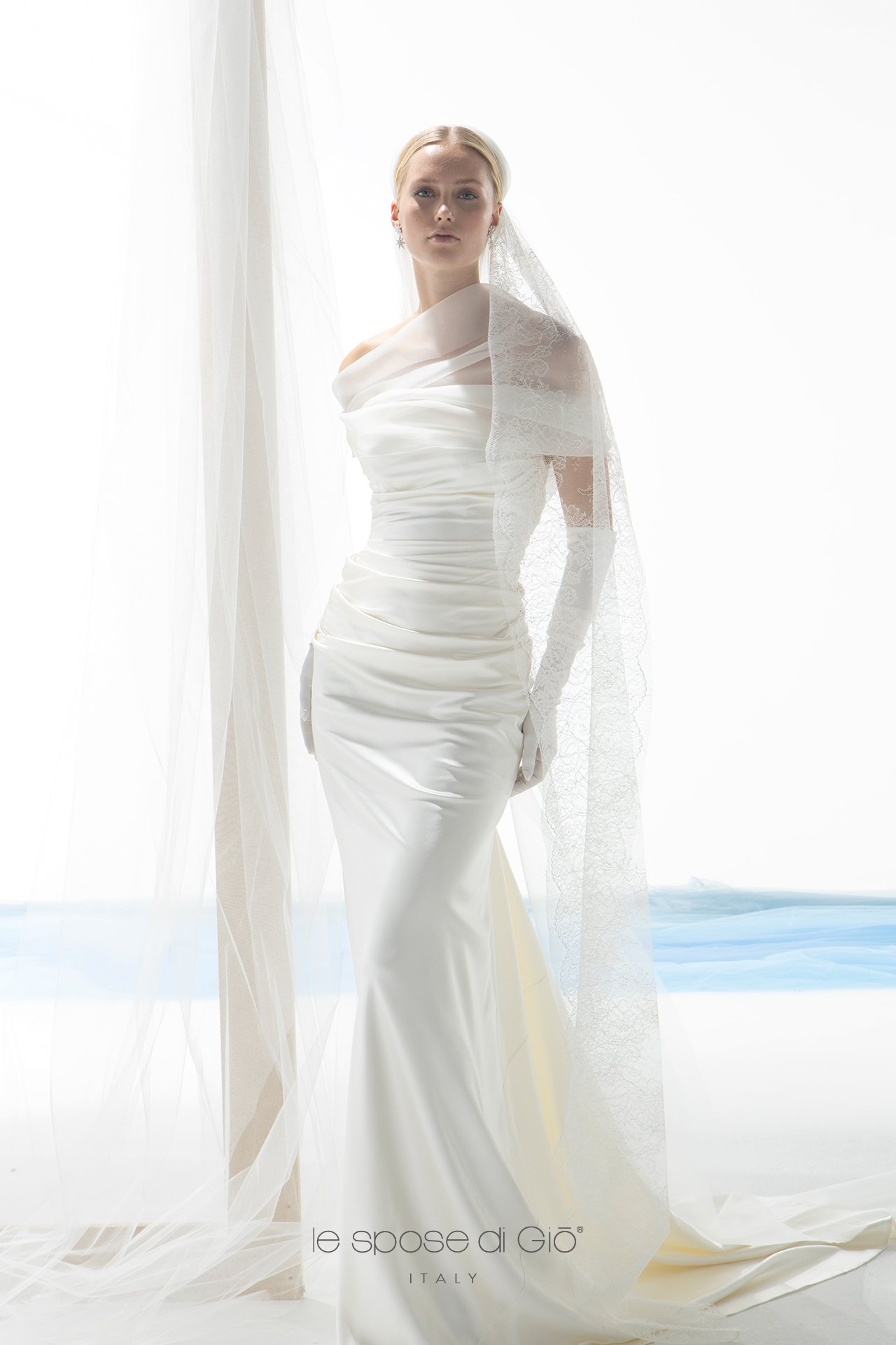 A woman in a white wedding dress with off-shoulder design, lace veil, and gloves stands in front of white curtains and bright lighting, with the logo 'le spose di Gio ITALY' at the bottom.