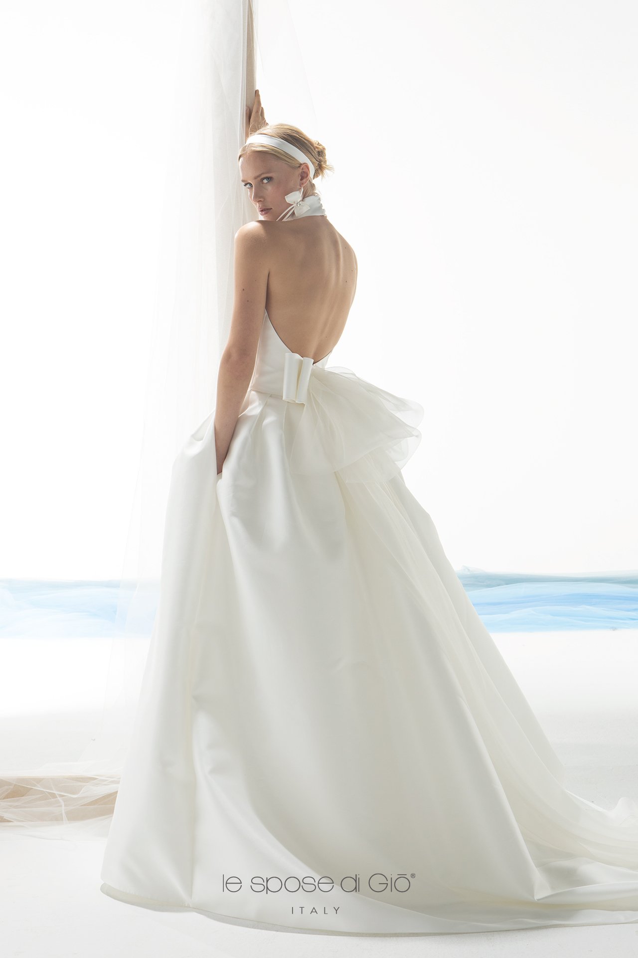 A woman in a white wedding gown with a full skirt, backless design, and bow detail, standing by a window with sheer curtains, looking over her shoulder.