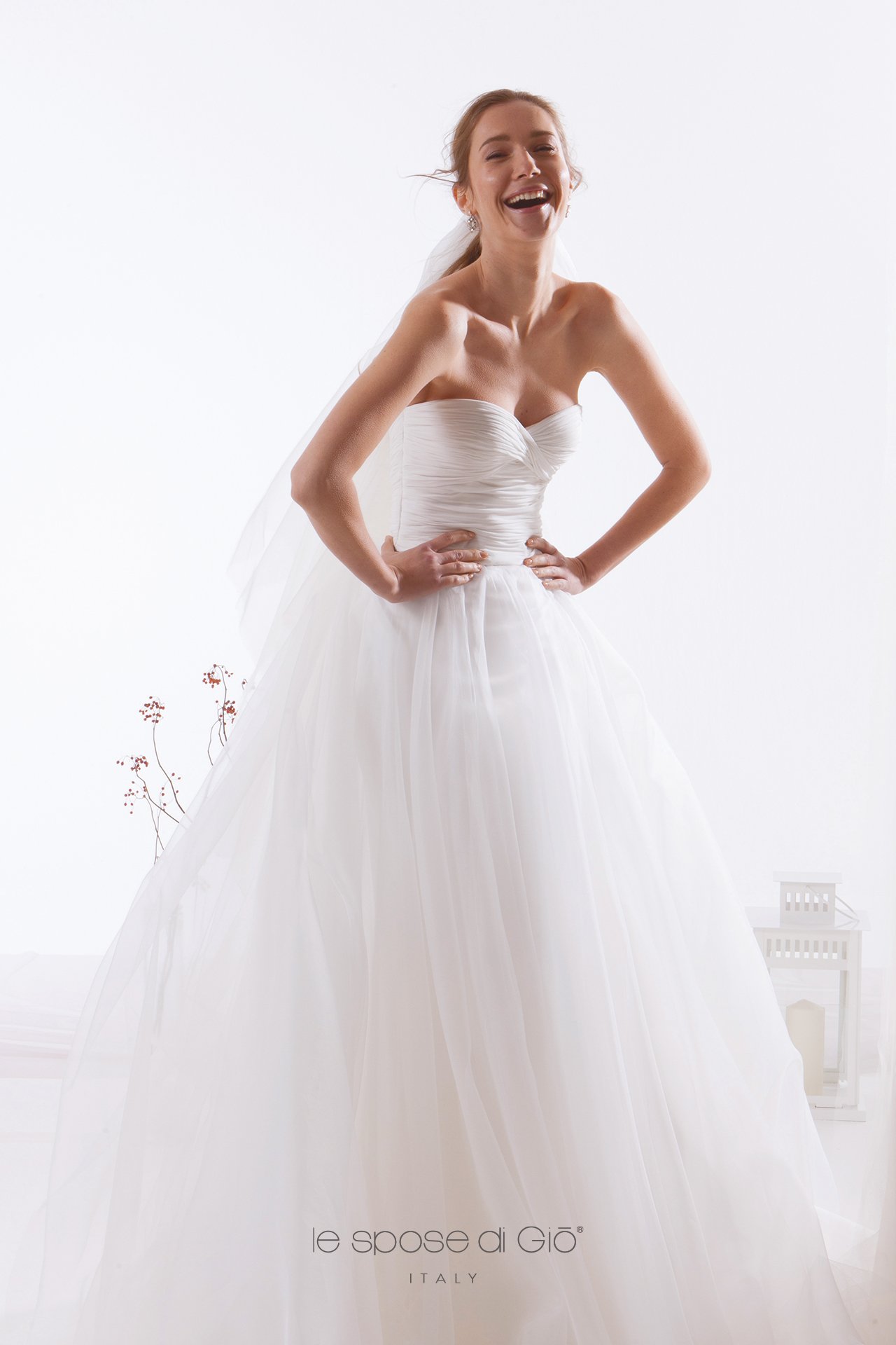 Smiling bride in strapless white wedding gown with full tulle skirt and veil, standing with hands on hips in a bright room.