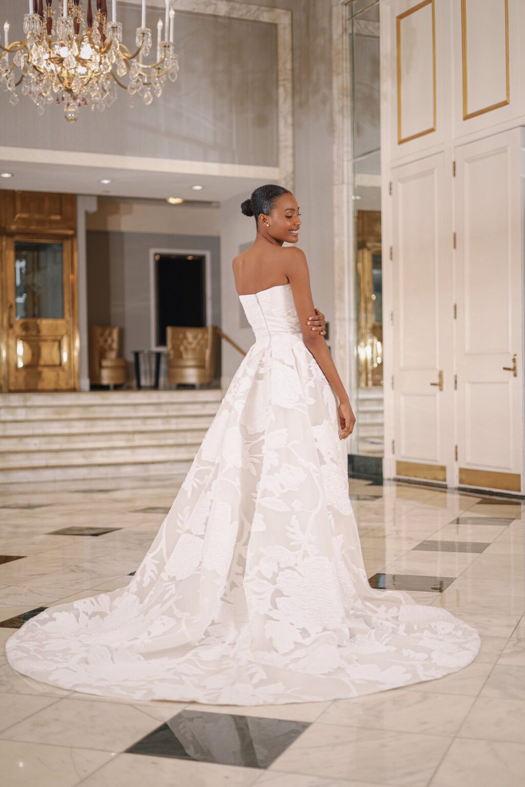 A woman in a strapless white wedding gown with floral patterns standing in a luxurious hall with marble floors, chandeliers, and gold accents.