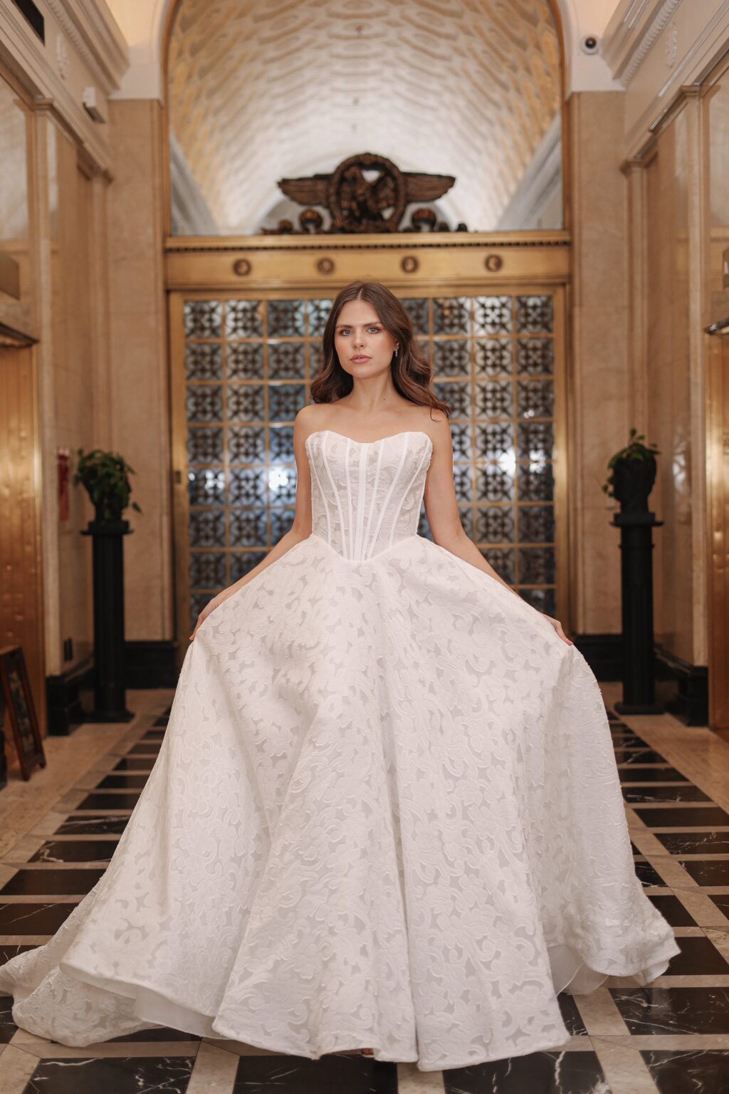 A young woman in a strapless white wedding gown standing in an ornate hall with marble floors and decorative walls, holding the sides of her full skirt.