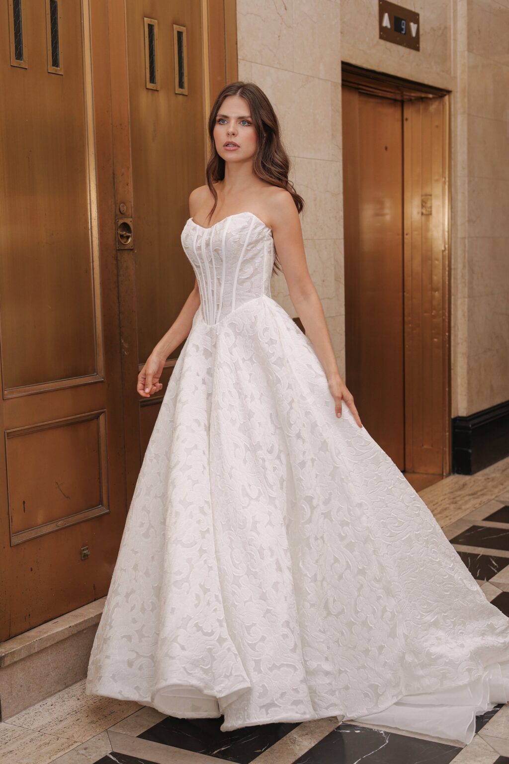A woman wearing a white wedding gown with a fitted bodice and a full skirt standing in an elevator lobby.