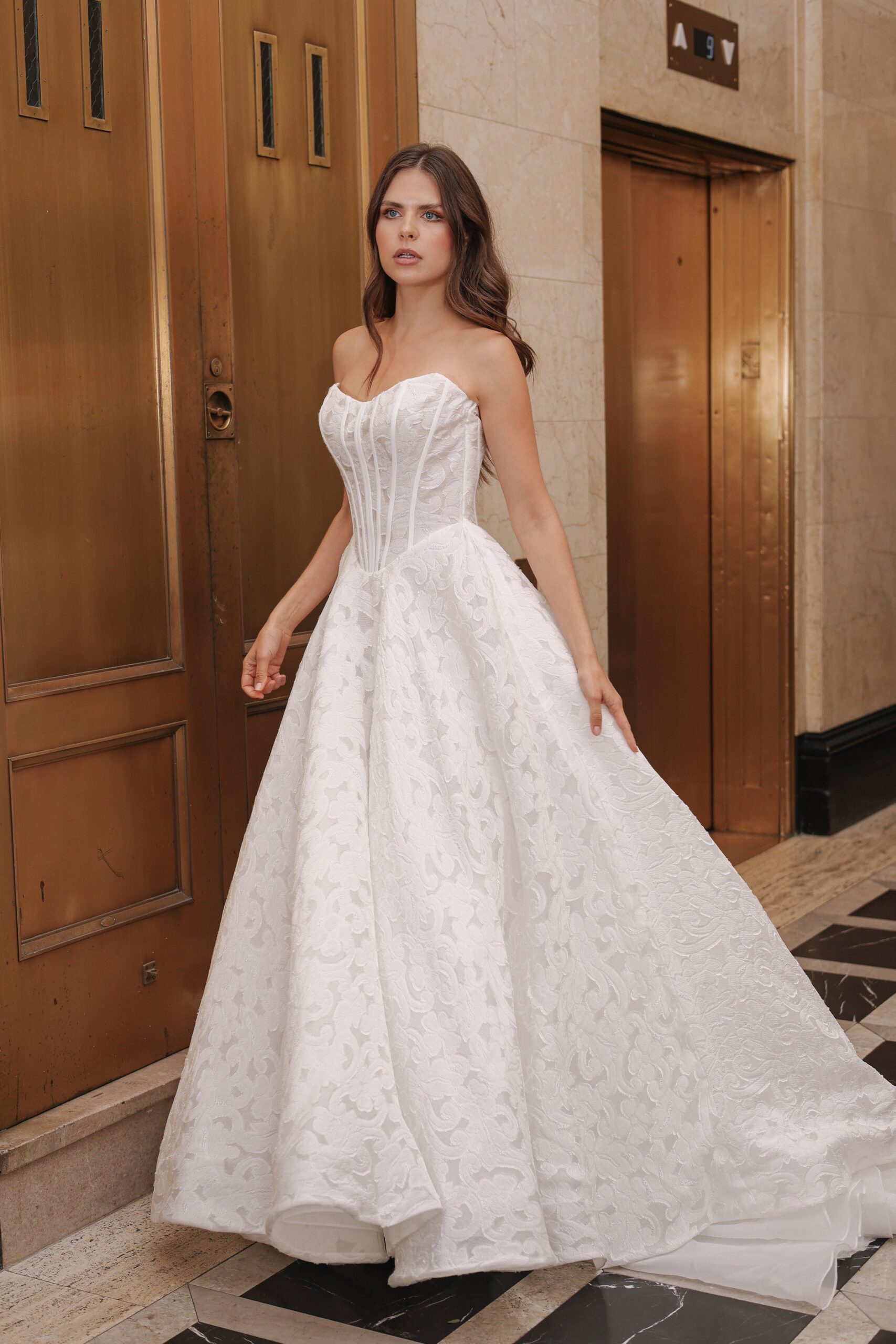 A woman wearing a white wedding gown with a fitted bodice and a full skirt standing in an elevator lobby.