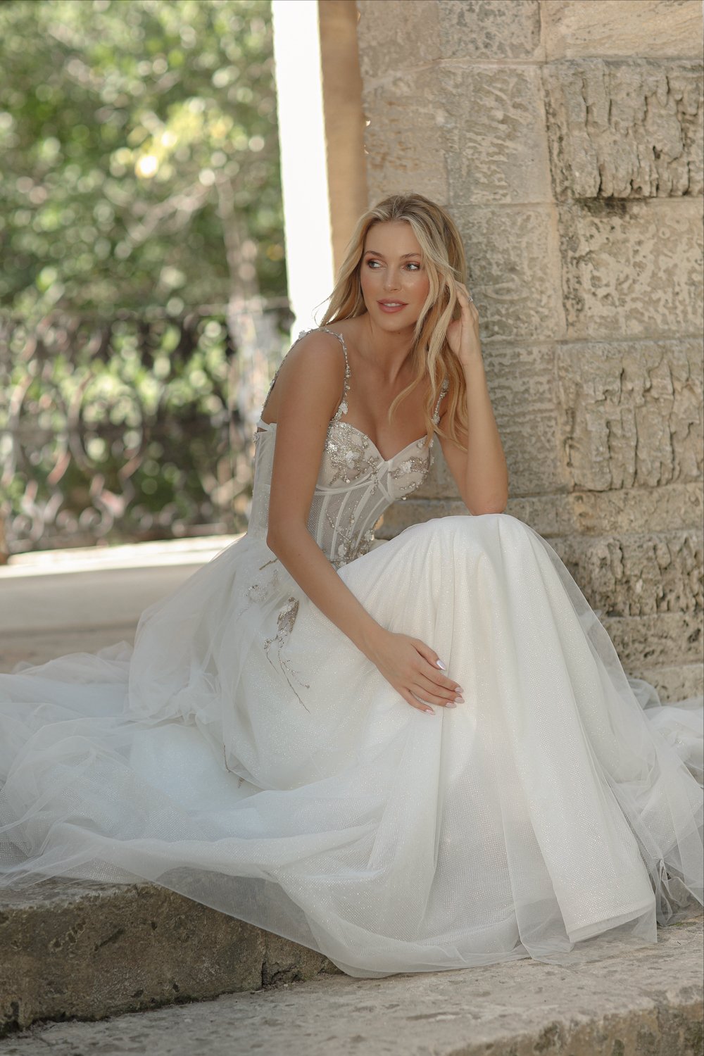A woman in a wedding dress sitting outdoors near a stone wall, with greenery in the background.