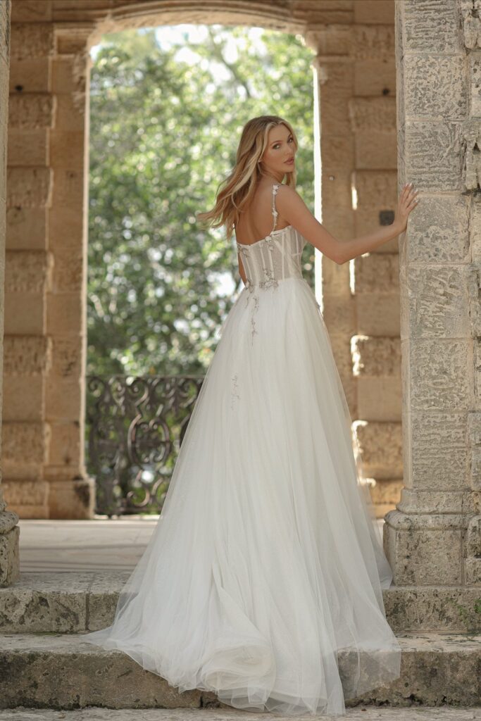 A woman in a white wedding dress standing on stone steps at a stone archway, with trees visible in the background.