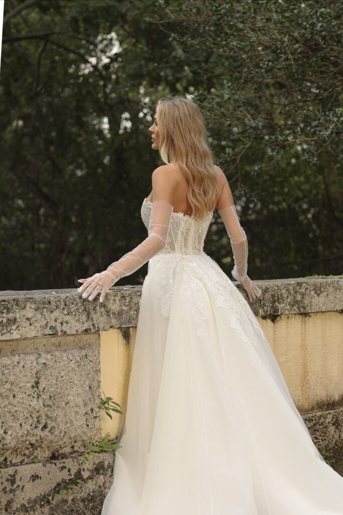 A woman in an elegant white wedding gown with lace and embroidery details, wearing sheer gloves, standing outdoors by a stone wall with trees in the background.