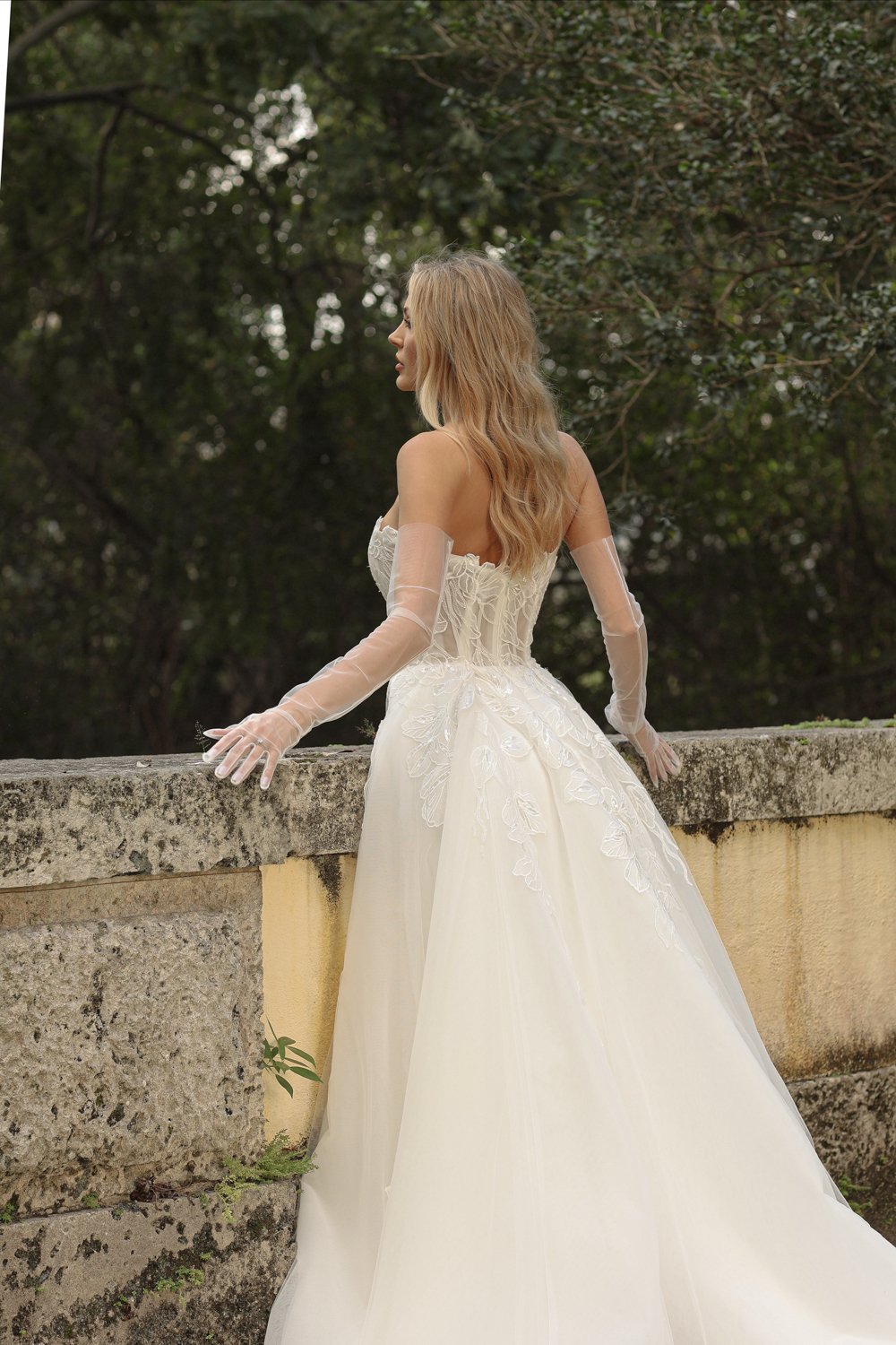 A woman in an elegant white wedding gown with lace and embroidery details, wearing sheer gloves, standing outdoors by a stone wall with trees in the background.