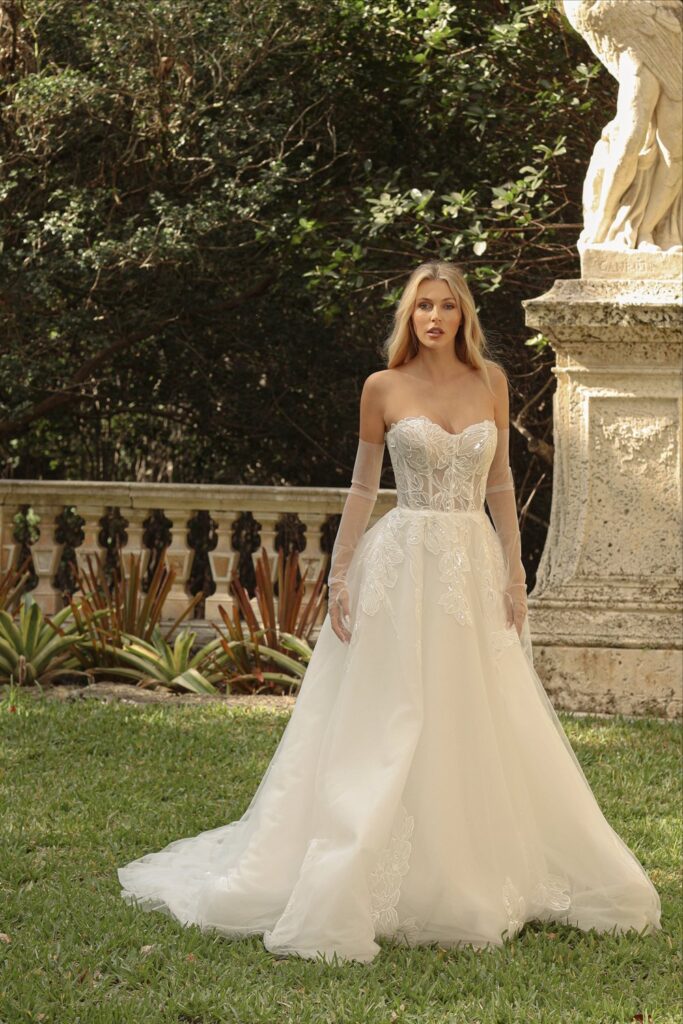 Woman in a white wedding dress standing outdoors on grass with trees and a classical sculpture in the background.