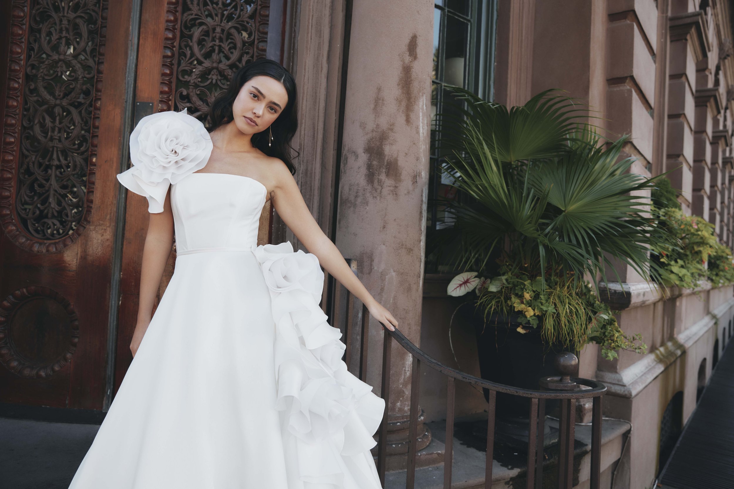 A woman in a white wedding dress with large floral adornments on her shoulder and waist standing outdoors beside a decorative iron railing and a large potted plant, with a brown building in the background.