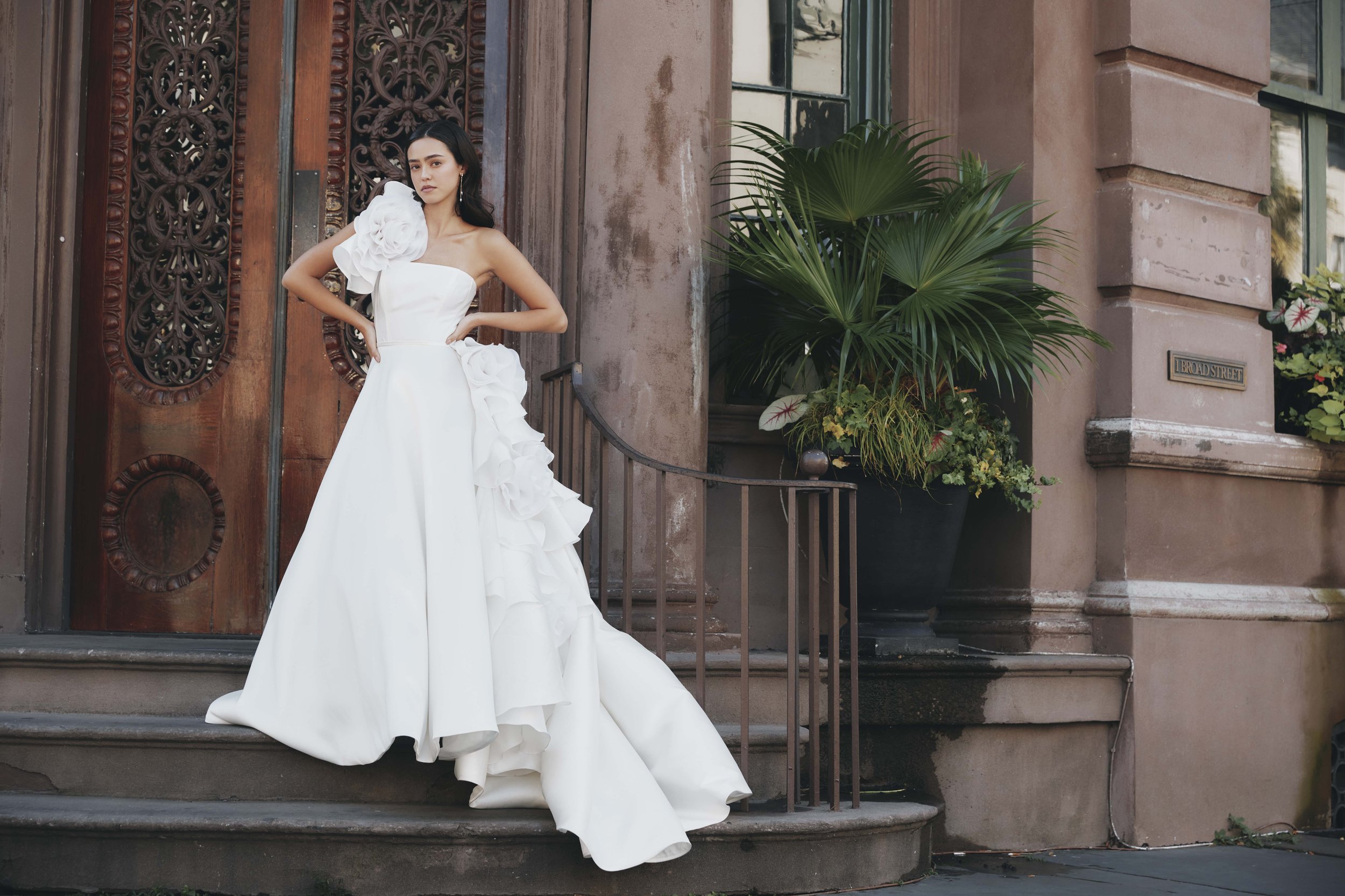 A woman in a white wedding gown with large floral embellishments stands on the front steps of a brownstone building, with one hand on her hip and the other on her waist, next to a large potted plant.