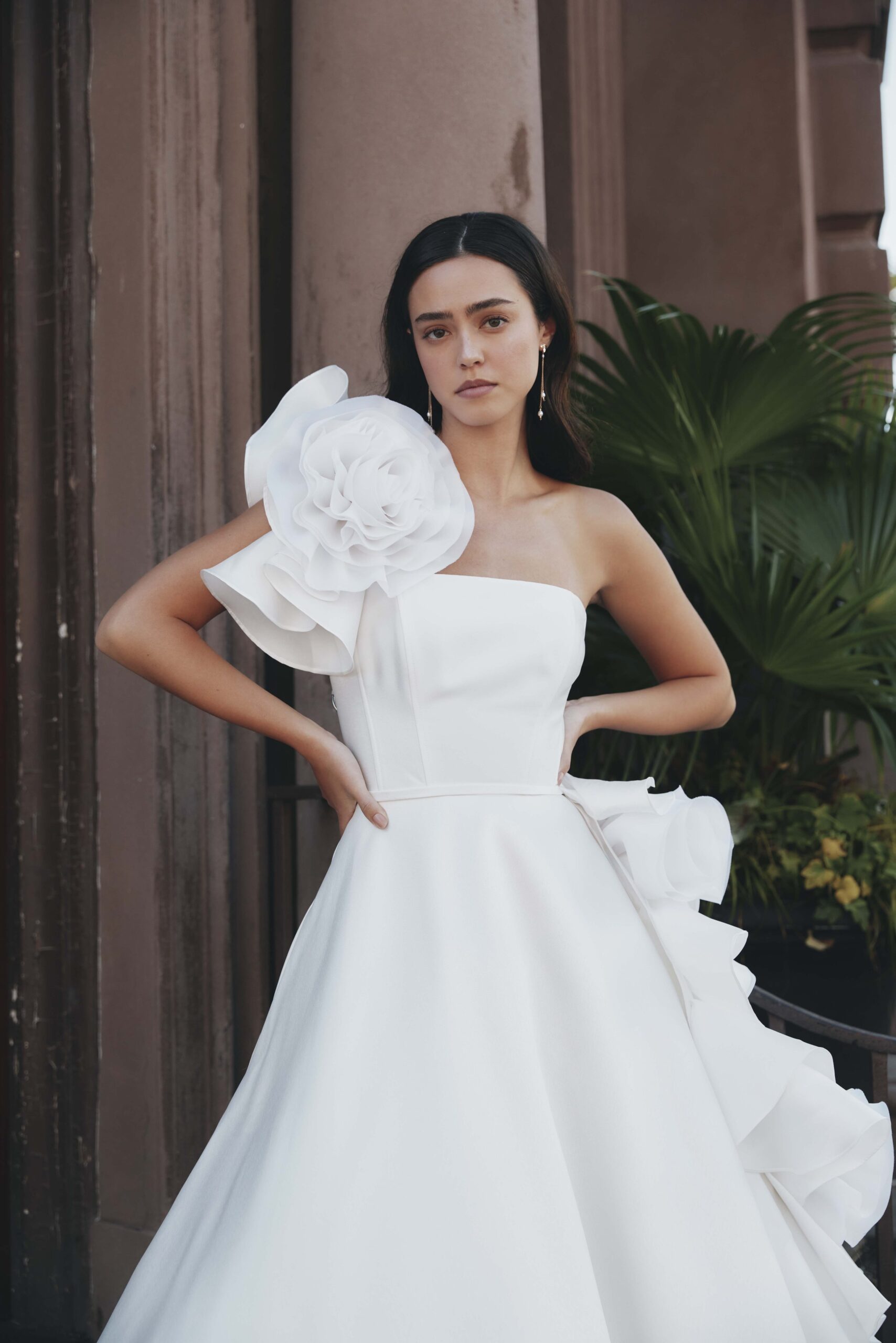 A woman wearing a white strapless gown with large floral embellishments on the shoulder and waist, standing outdoors near a building with green plants in the background.