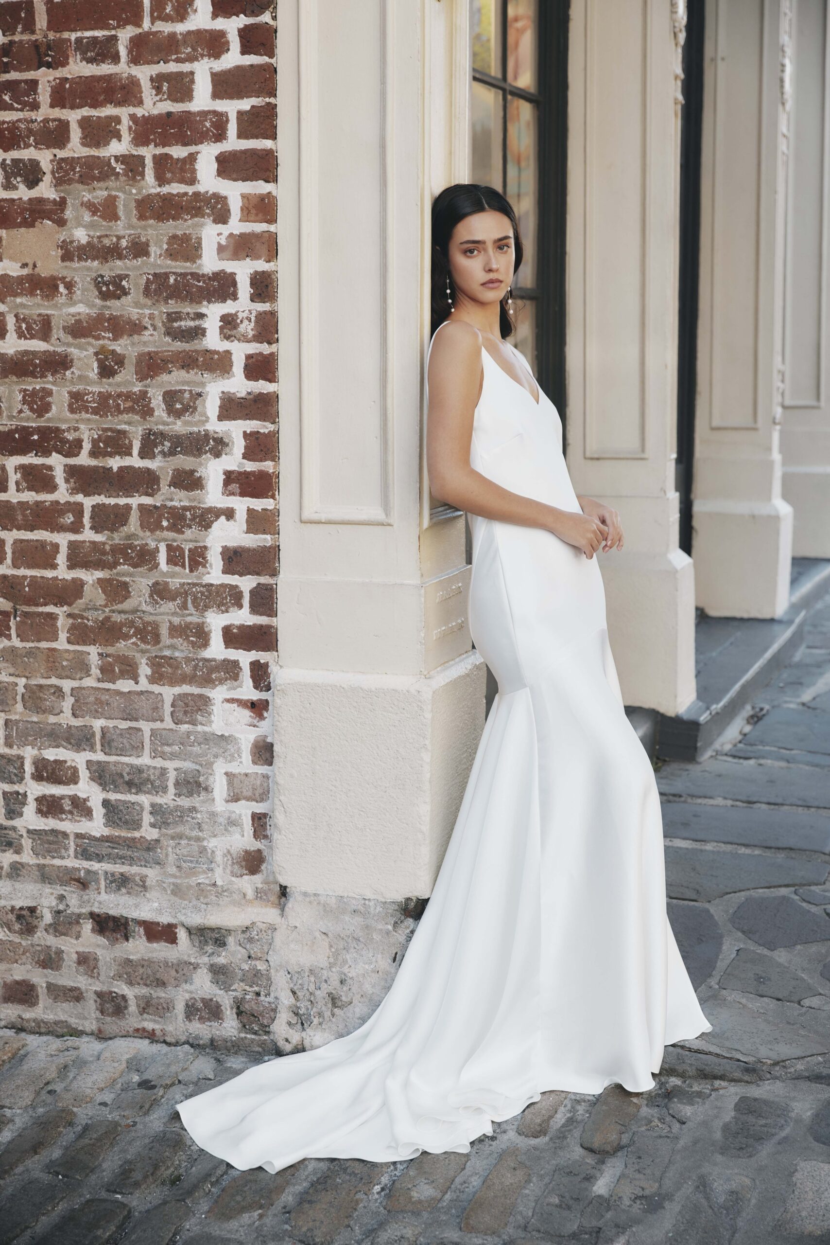 Woman in a white wedding dress leaning against a white wall outside a brick building.