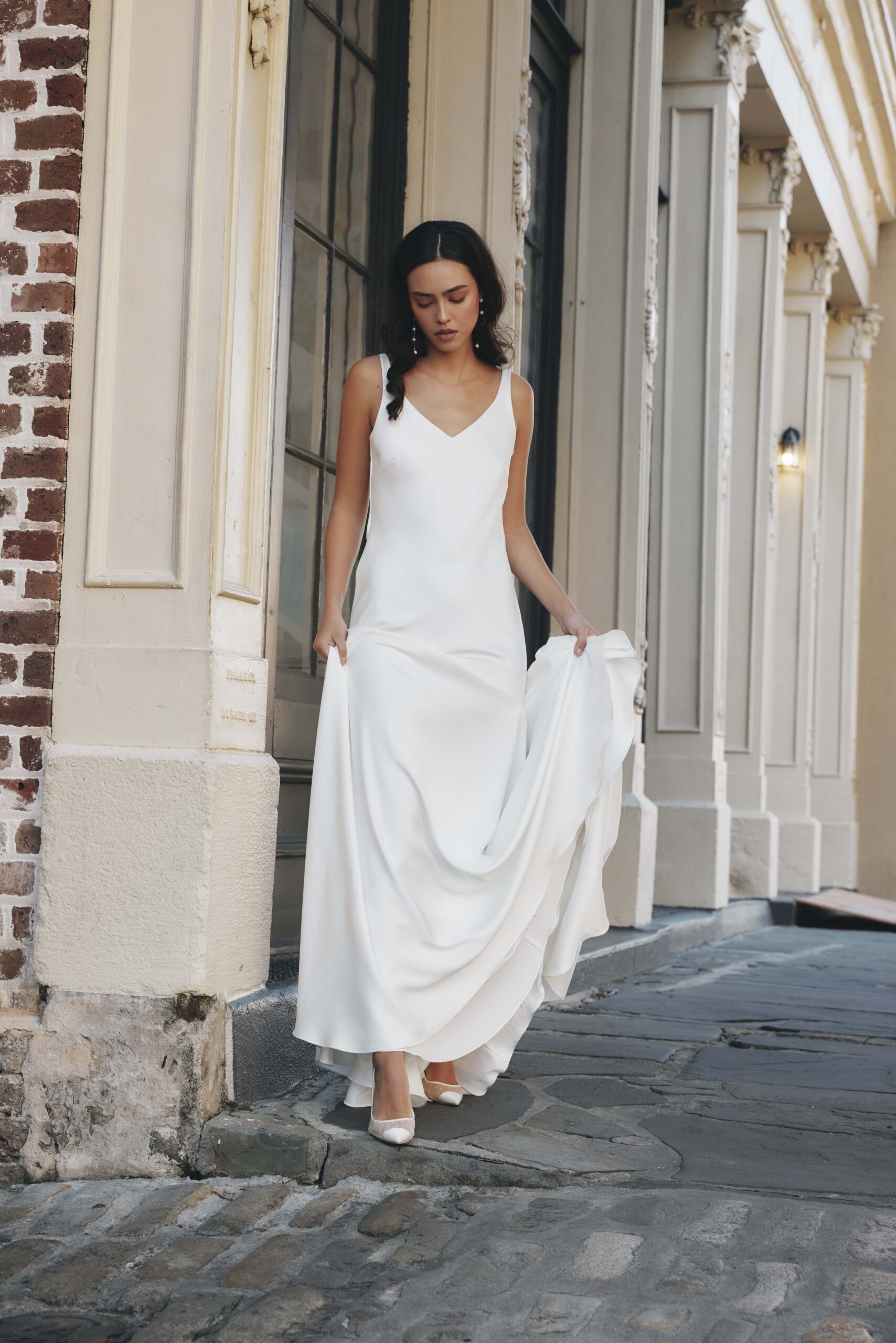 A woman in a long white dress walking on cobblestone street outside of a building with decorative white trim and brick accents.