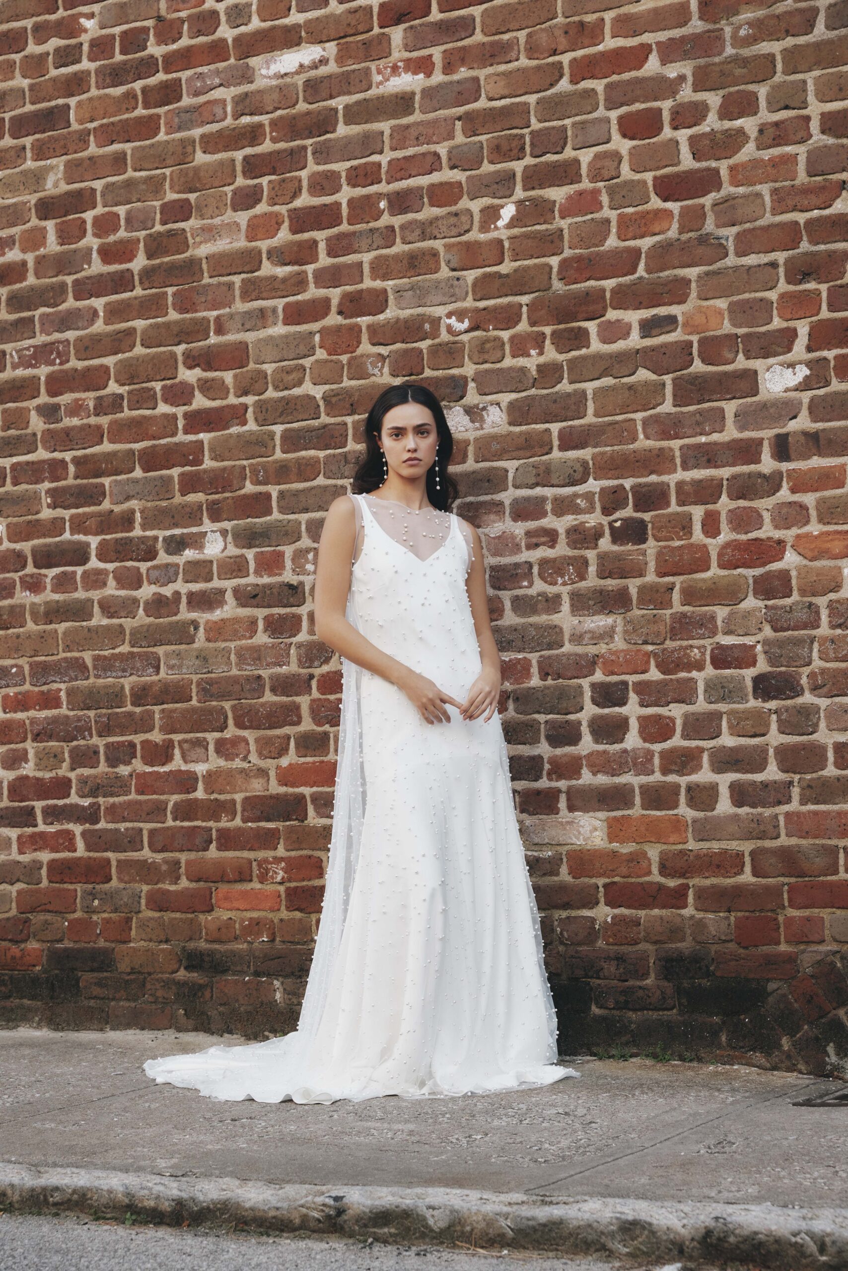 A woman in a white dress with pearl details standing against a brick wall.