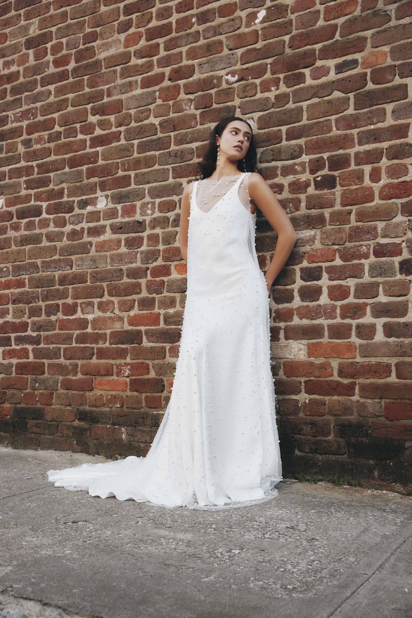 A woman in a white dress with pearl embellishments, standing against a brick wall, looking to her left with her hands in her pockets.
