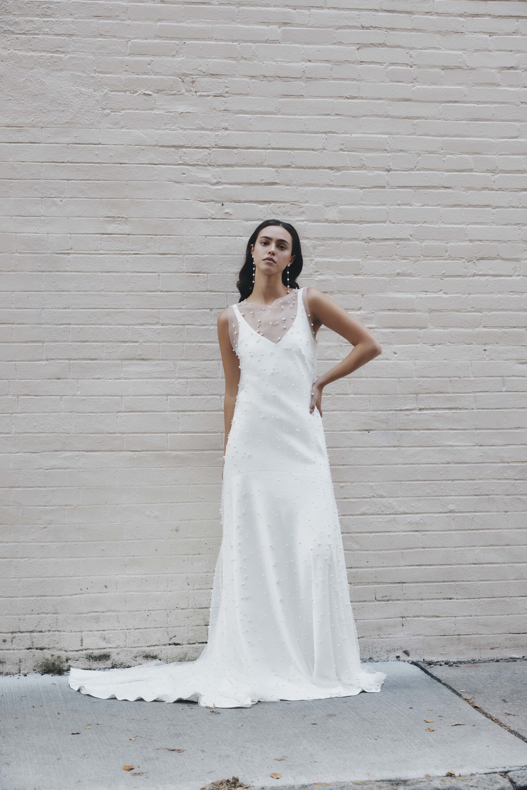 A woman posing in a white dress with pearl embellishments, standing against a beige brick wall.