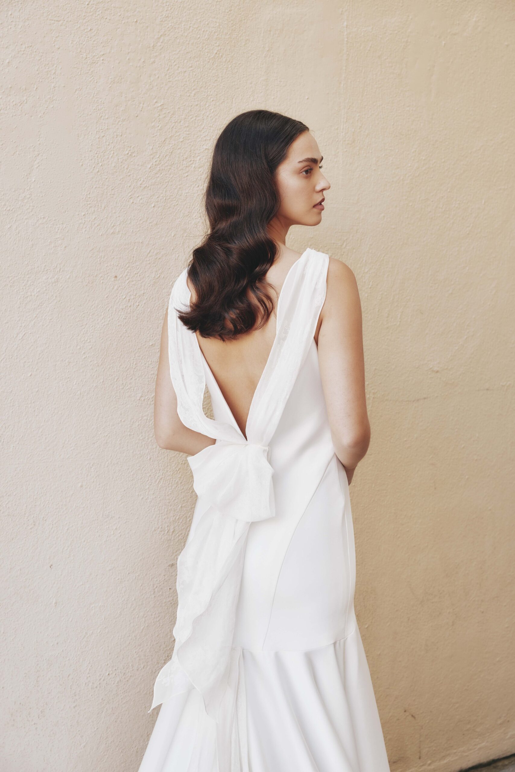 A woman with wavy brown hair wearing a white dress with an open back and bow detail, standing sideways against a beige wall.