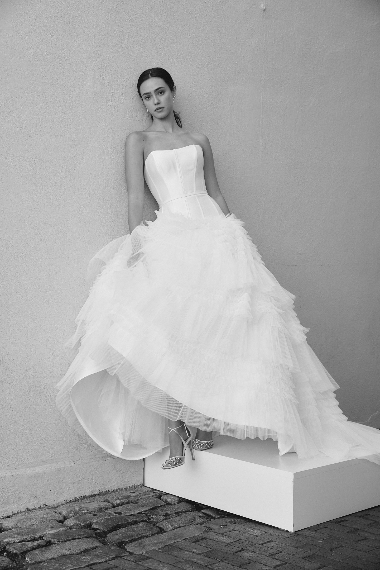 A woman in a strapless wedding gown with a fitted bodice and full layered tulle skirt stands on a white platform against a textured wall. She wears strappy high heels and has dark hair styled back.