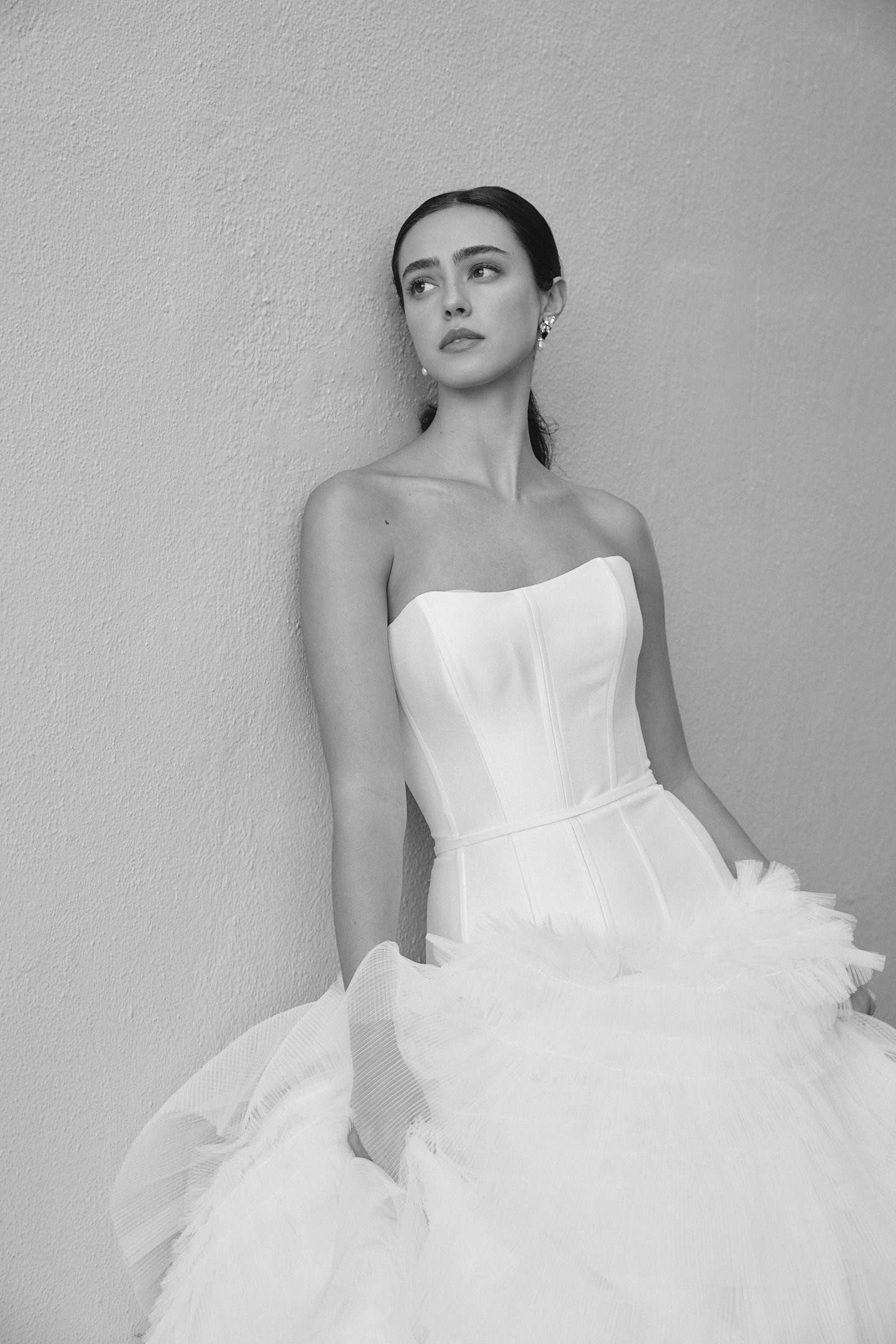 A woman in a strapless white dress with a ruffled skirt, standing against a plain wall, looking to the side.