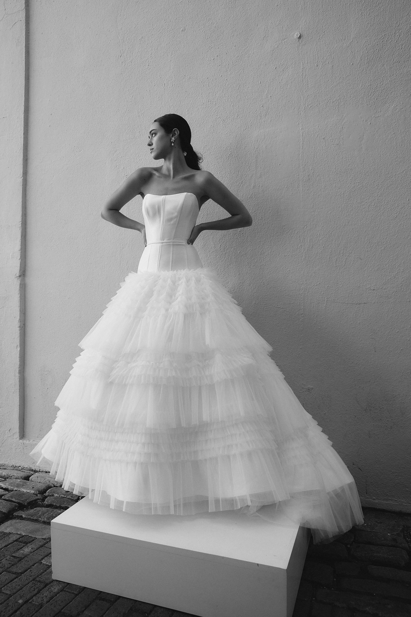 A woman in a strapless white wedding gown with a tiered tulle skirt stands on a white platform against a plain wall.