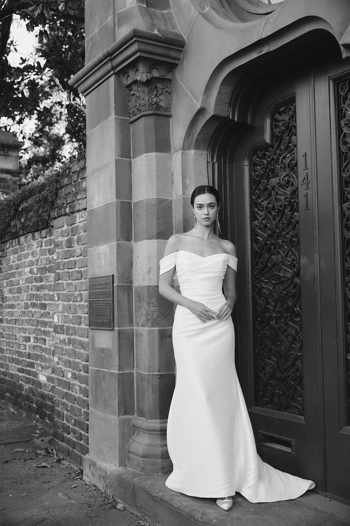 A woman in an elegant off-the-shoulder white dress standing outside a historic building with ornate dark door and brick wall.