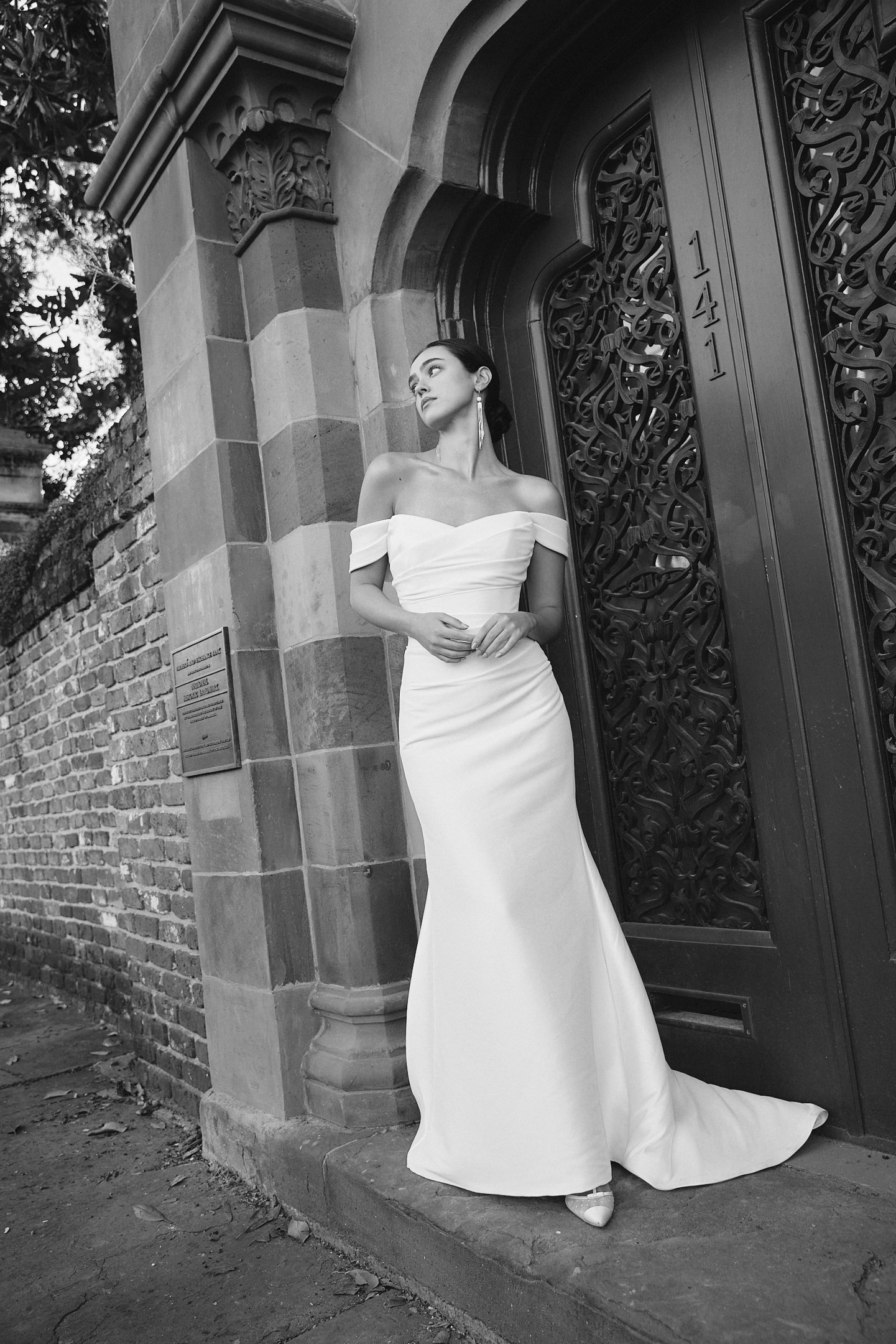 A woman in an off-shoulder white wedding gown standing beside an ornate entrance with a large arched door, decorative ironwork, and brick and stone architecture. She is looking to the side with her hands clasped in front of her.