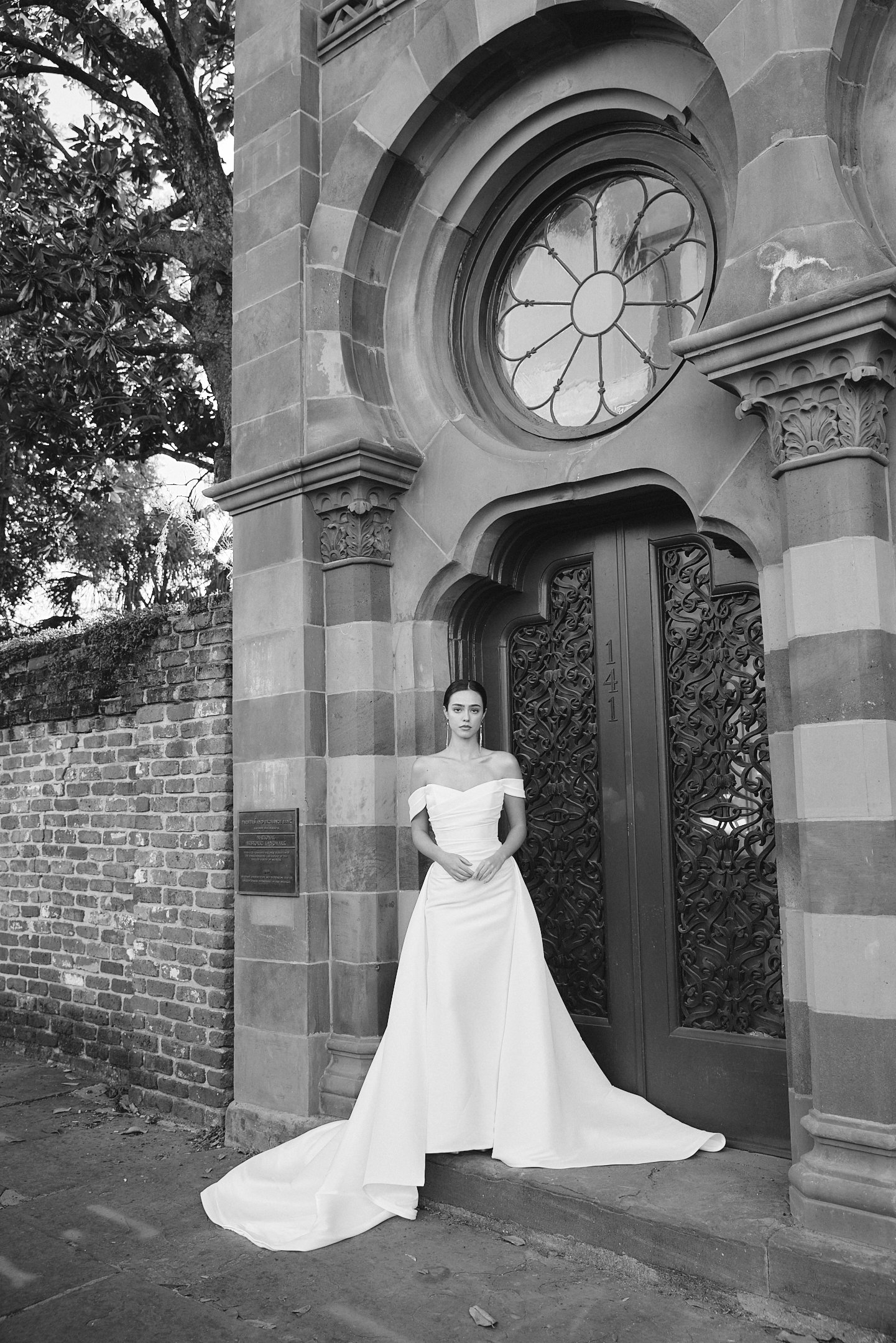 A woman in a wedding dress standing in front of an ornate, arched entrance with decorative ironwork and stained glass window, with a brick wall and trees in the background.