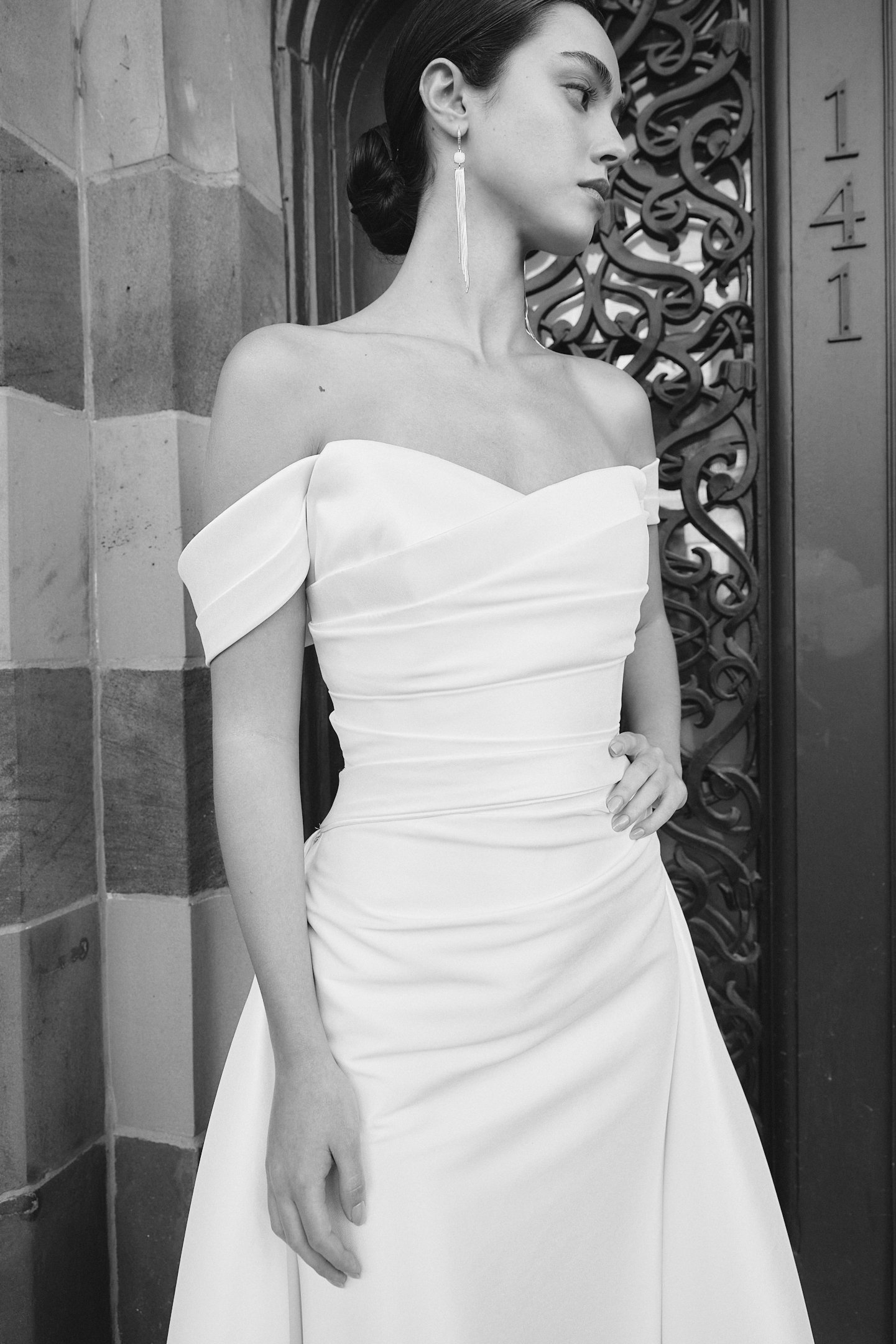 A woman in a white off-the-shoulder dress with a hand on her hip, silver dangling earrings, and her hair in an elegant bun, standing near an ornate door with the number 1411 on it, in black and white.