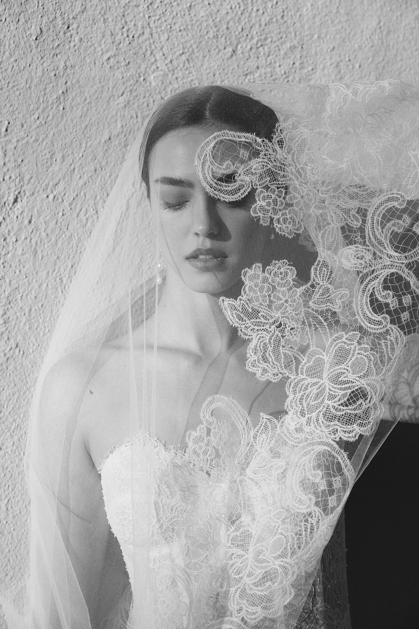 A woman in a wedding dress with lace accents and a veil, standing against a textured wall with her eyes closed.