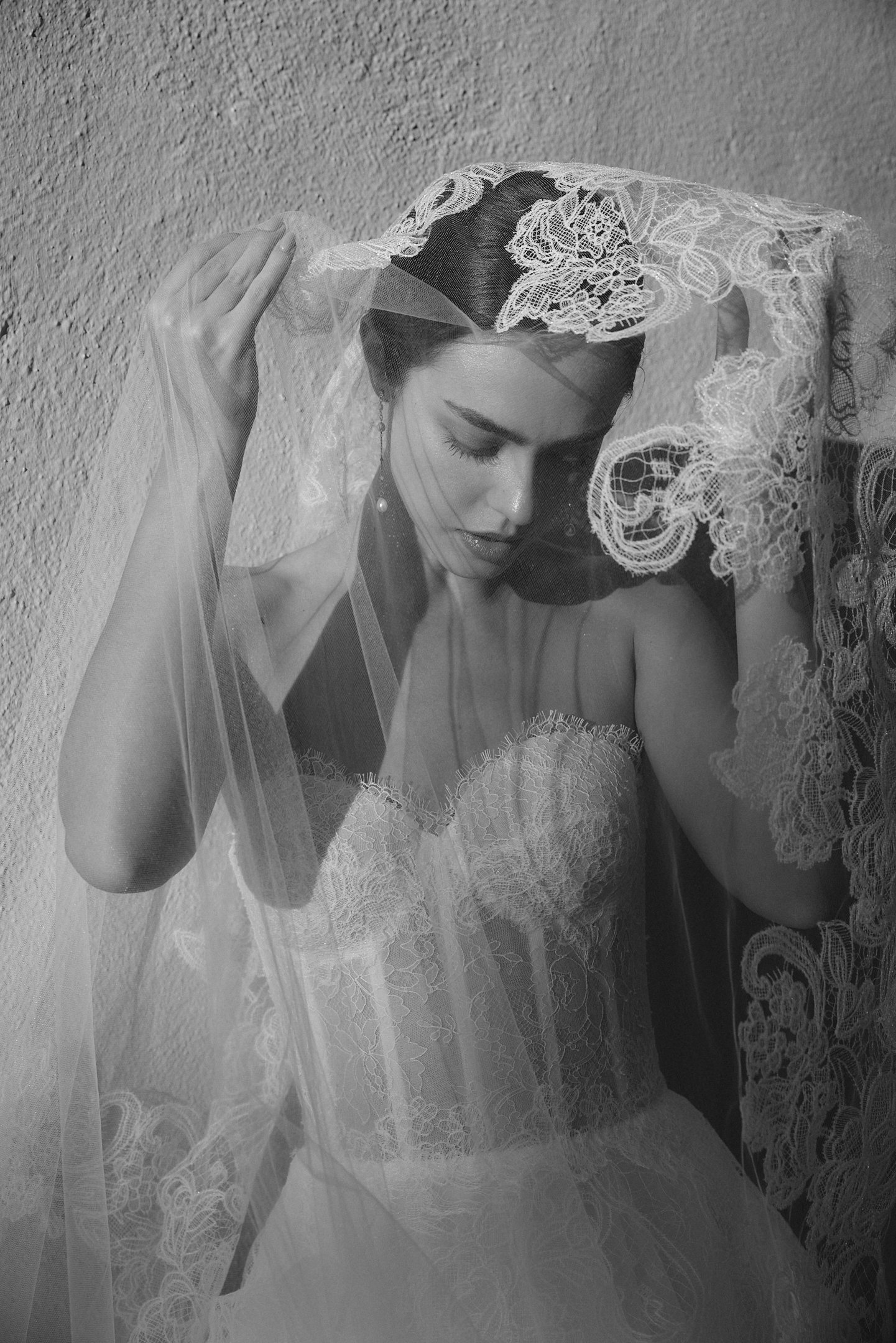 Black and white photo of a woman wearing a wedding dress and veil, with her eyes closed and head bowed, against a textured wall.
