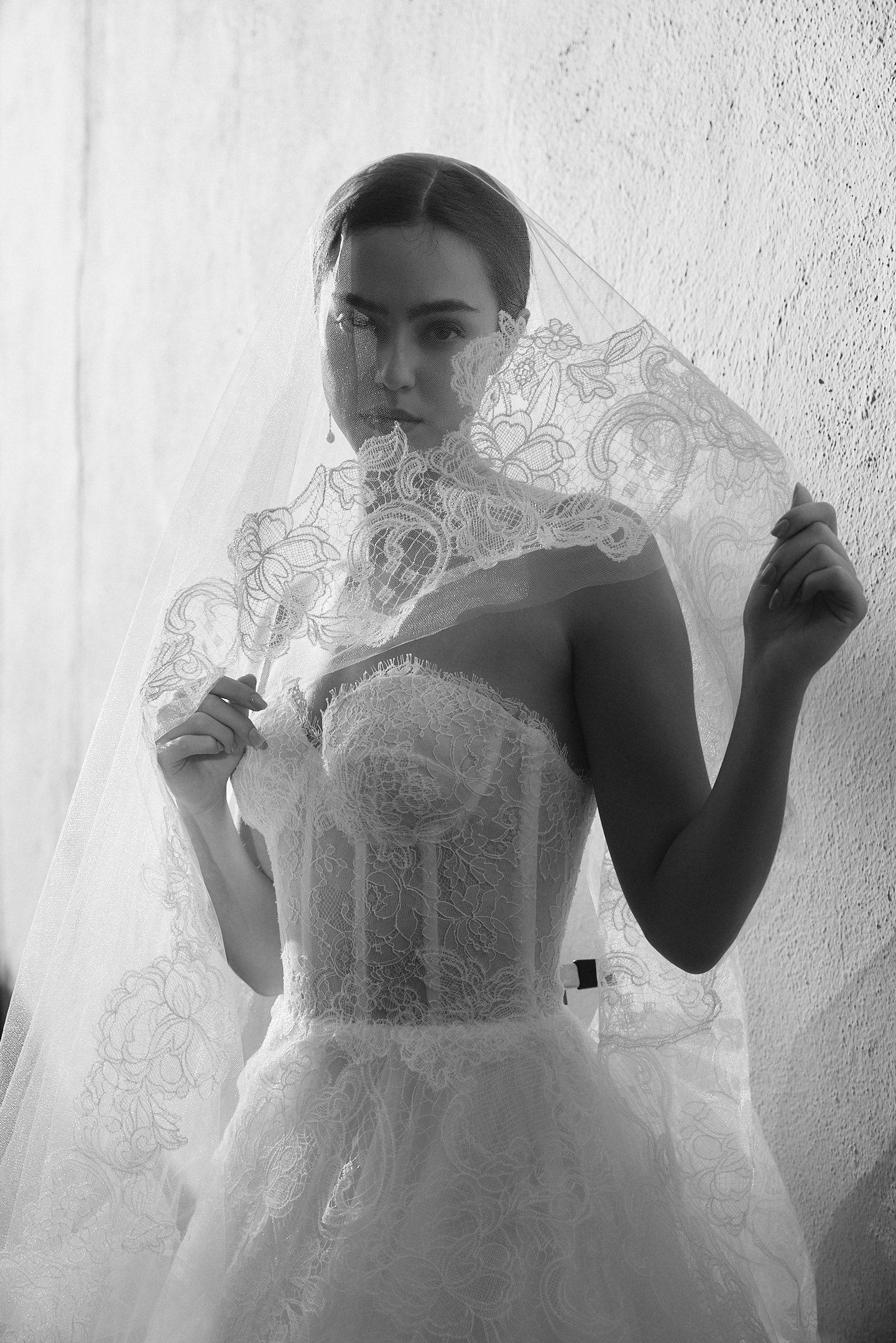 A woman in a wedding dress holding a lace veil in front of her face, standing against a textured wall.