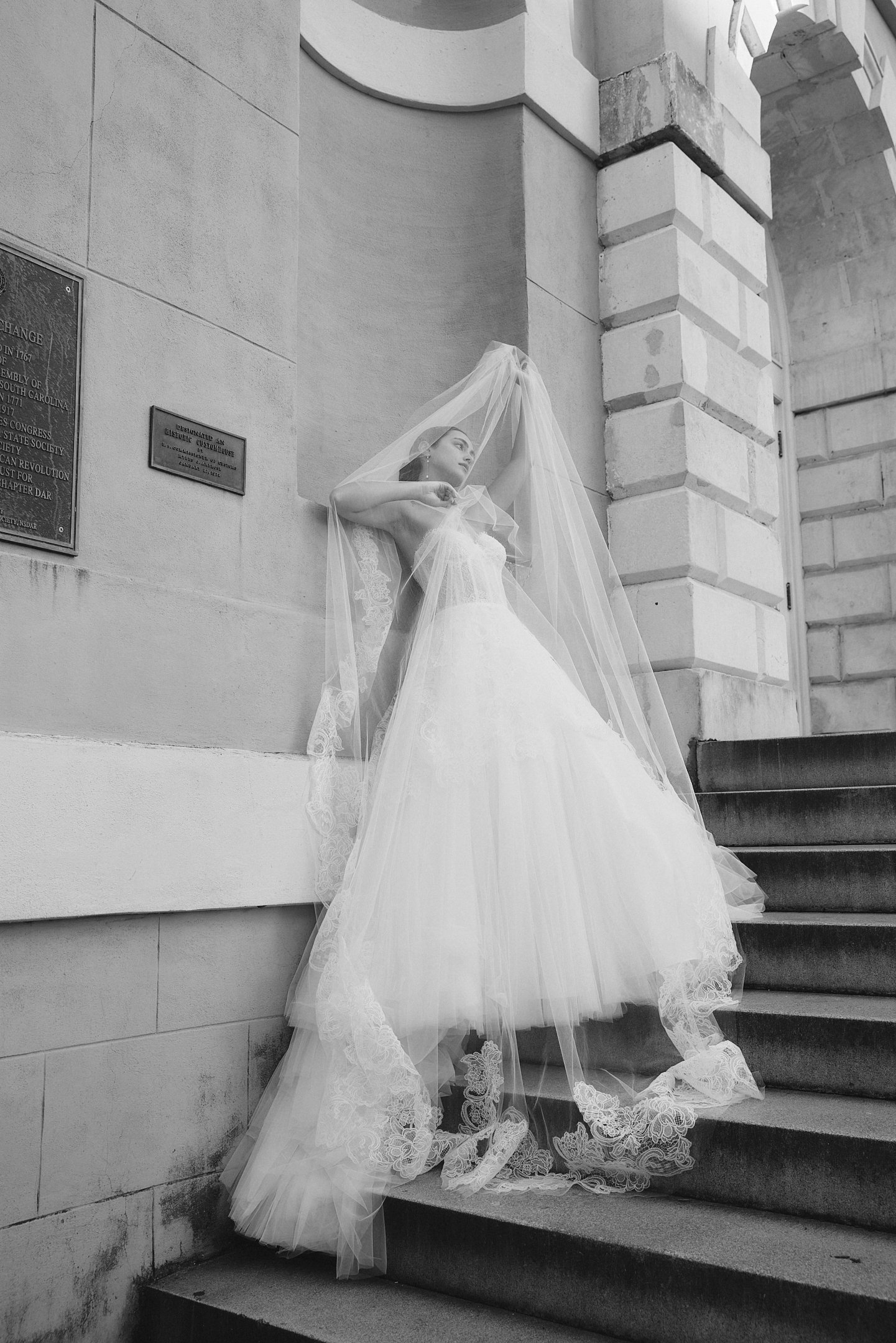 Black and white photo of a woman in a wedding dress and veil standing on staircase, leaning against a stone wall with plaques, with her arms raised and head tilted back.