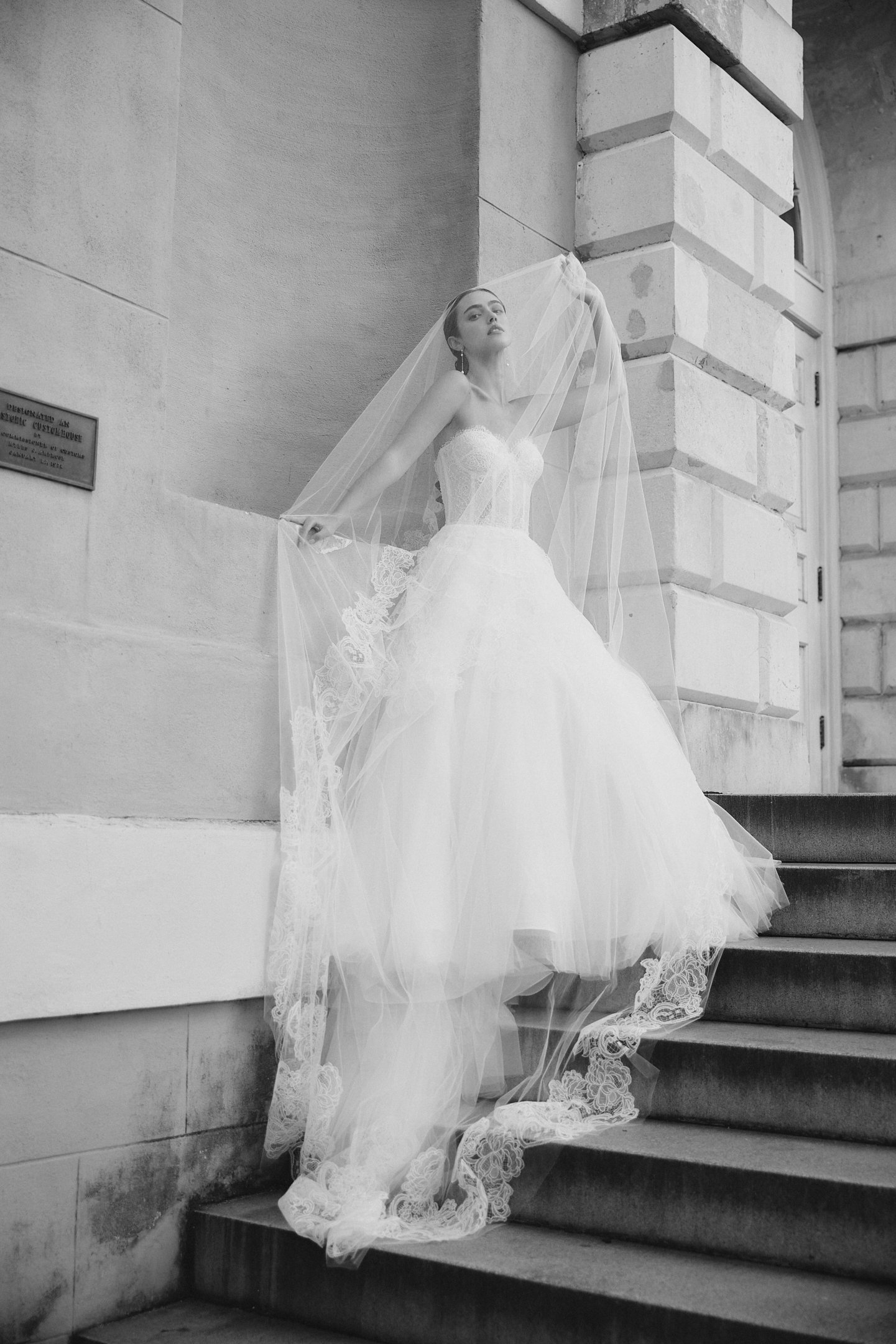 A woman in a wedding dress and veil stands on stairs outside a stone building, striking a pose with her hand holding her veil and looking into the distance.