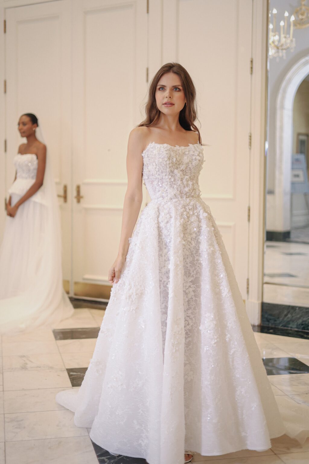 Two women in wedding dresses standing in an elegant room with white walls and marble floors, one in the foreground looking to the side and another in the background facing forward.