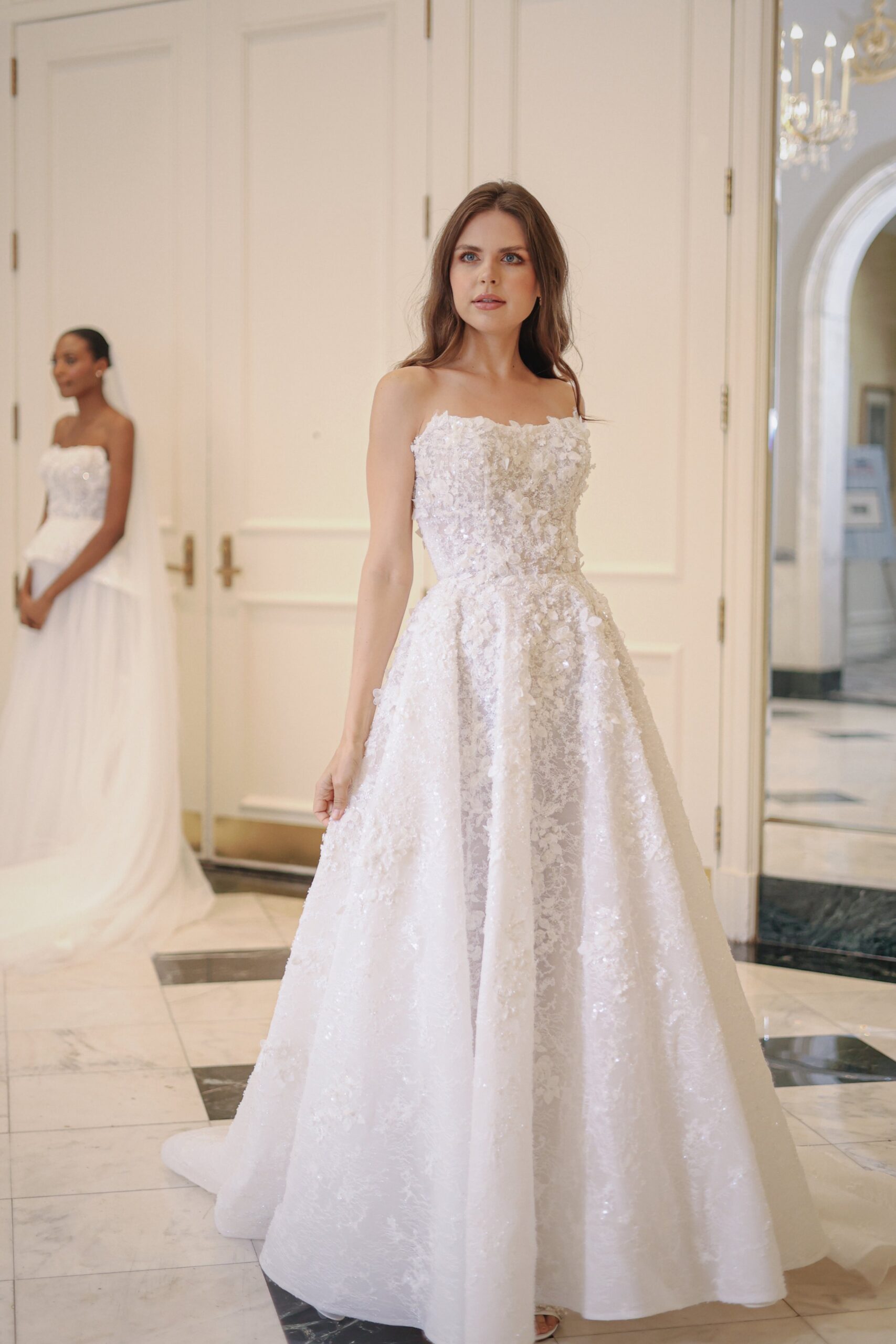 Two women in wedding dresses standing in an elegant room with white walls and marble floors, one in the foreground looking to the side and another in the background facing forward.