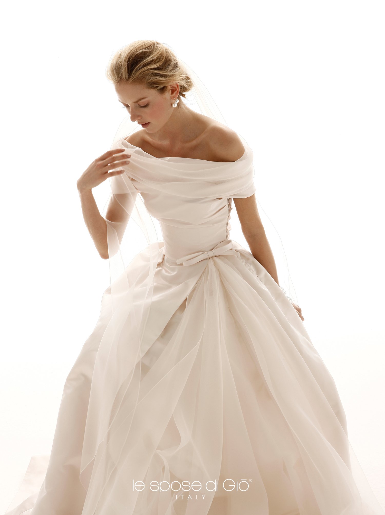 A woman in a flowing, off-shoulder, white wedding gown with sheer overlays, looking down and touching her shoulder, against a plain white background.