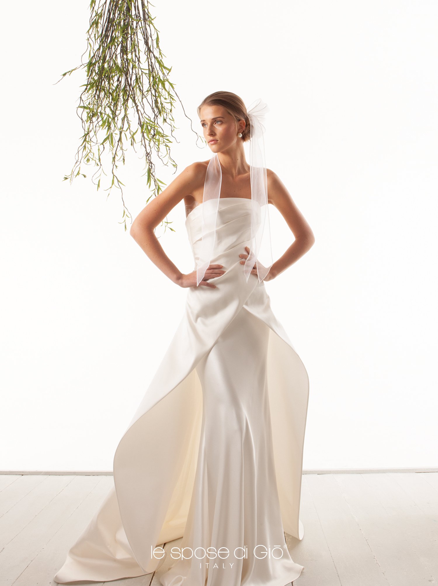 A woman in a white bridal gown standing with hands on hips, with hanging plant branches in the background and a mostly white setting.