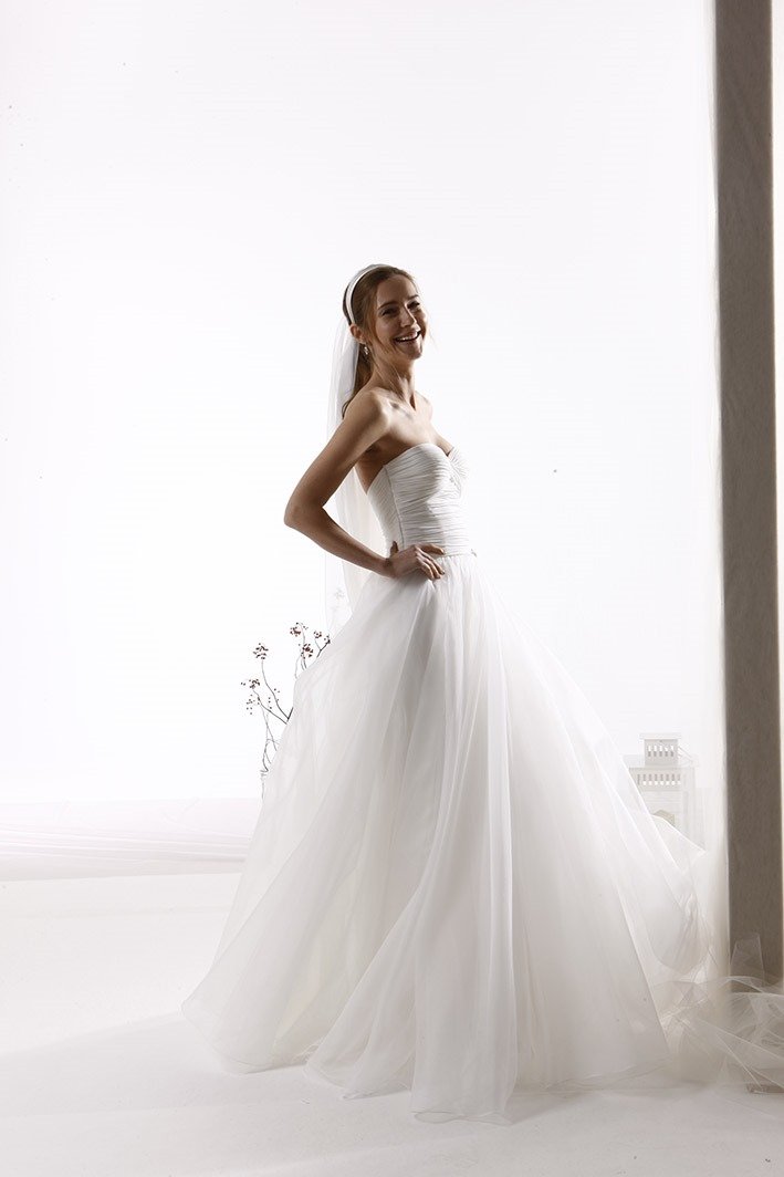 A smiling bride in a strapless white wedding gown with a full tulle skirt, standing indoors near a window with natural light.