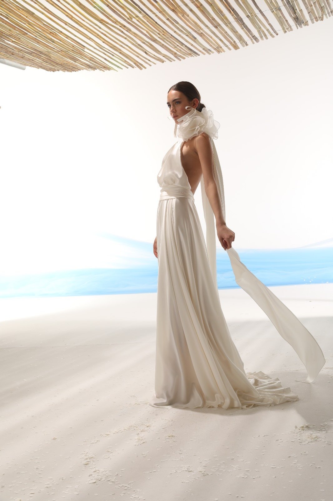 A woman in an elegant white gown with a large ruffled collar, standing on a sandy beach set with a backdrop of the ocean and sky, under a bamboo roof.