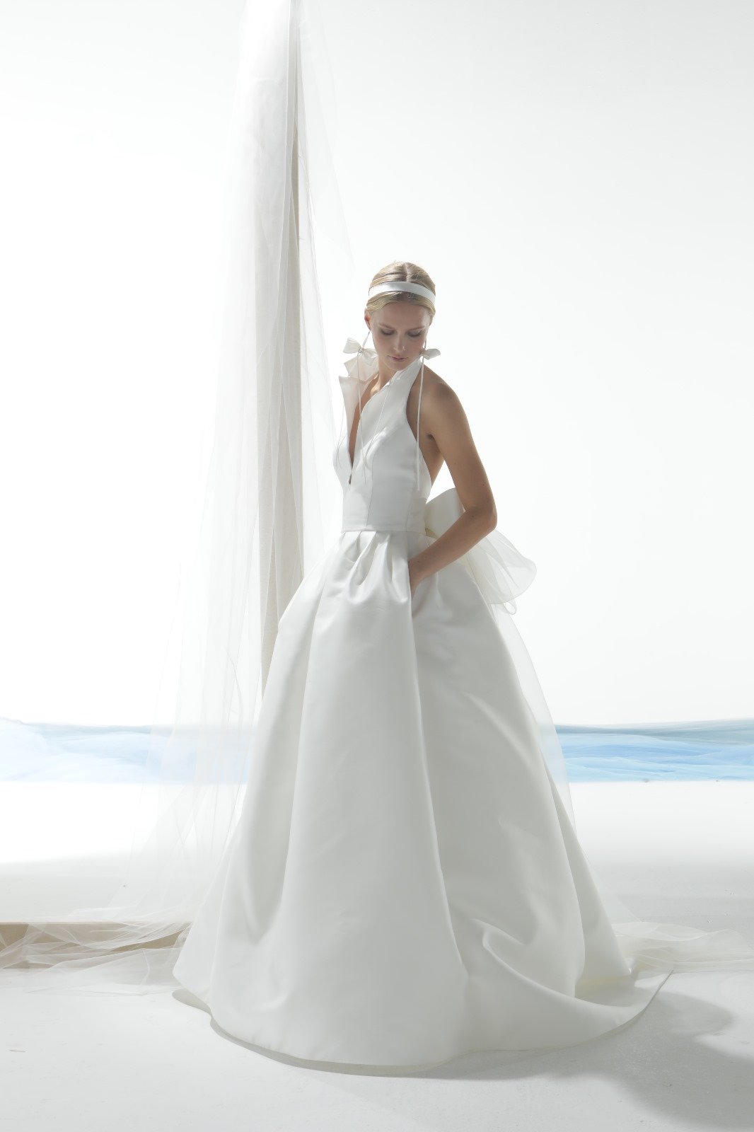 A woman in a strapless white wedding gown with a full skirt and pockets, standing in front of sheer white curtains, with her hair styled back and a white headband, looking down.