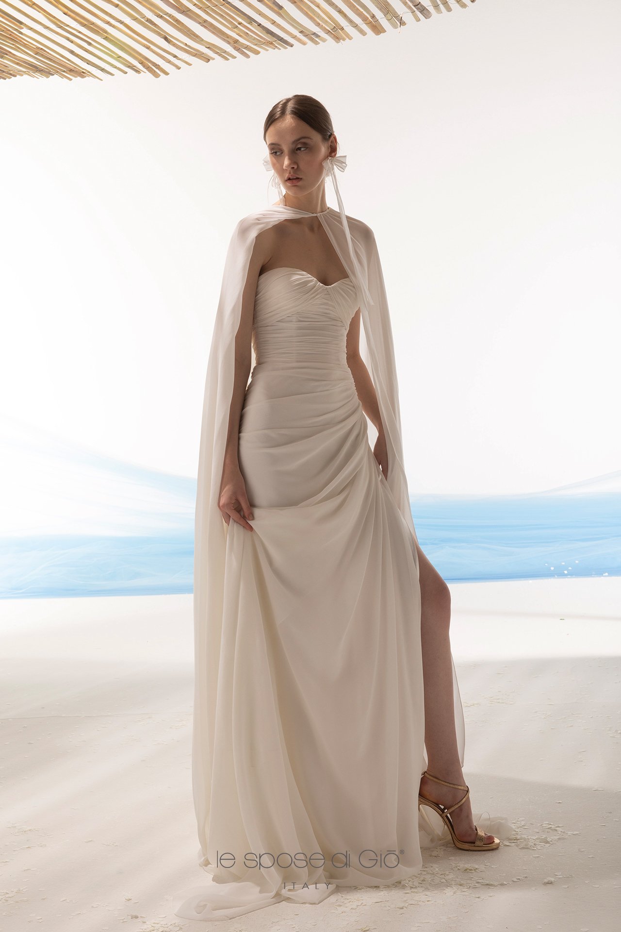 A woman in a white, strapless gown with ruched details and a thigh-high slit, standing on sand under a thatched roof with the ocean in the background.